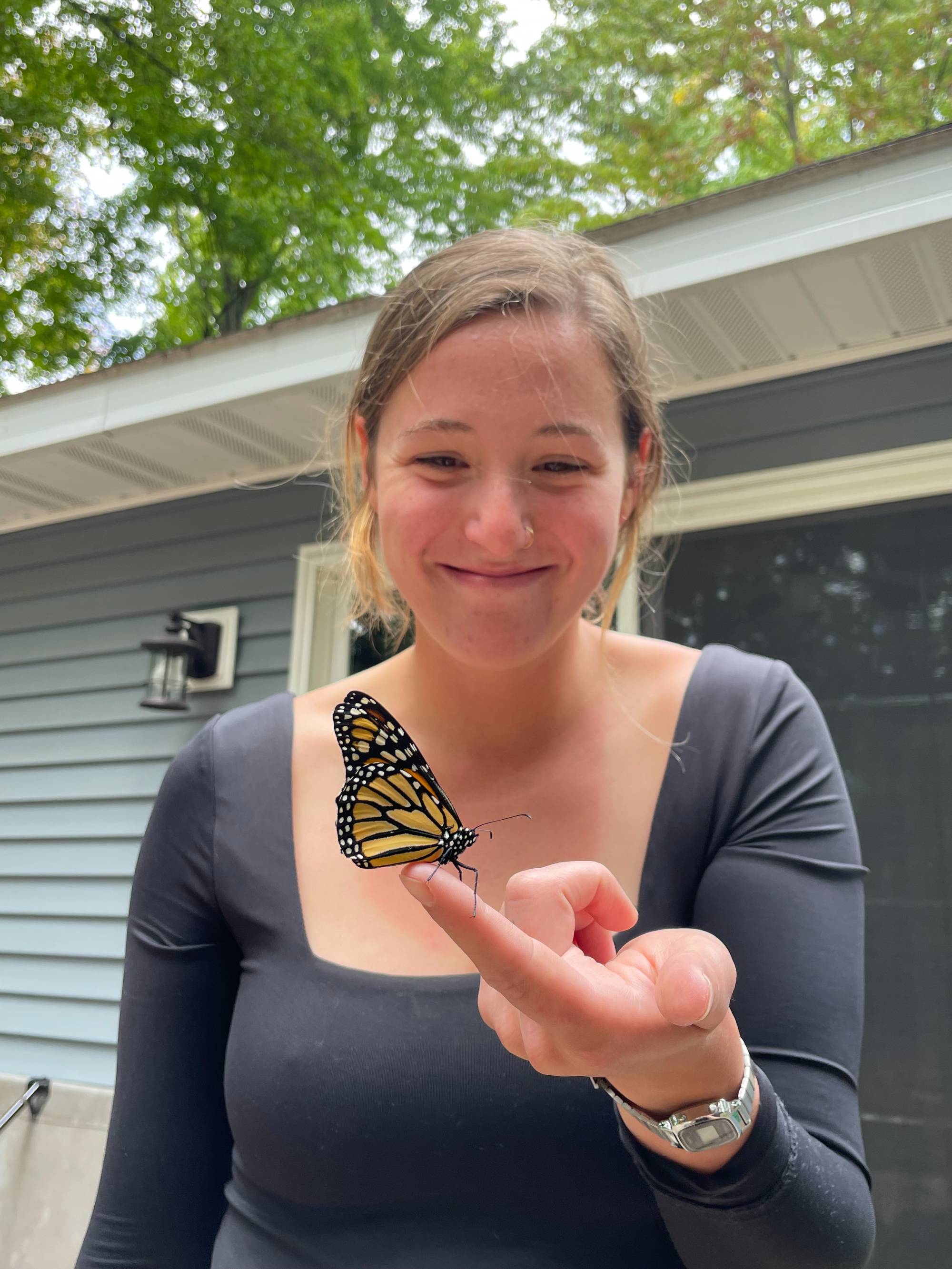 girl with brown hair wearing a grey shirt smiling softly while holding a monarch butterfly on her finger